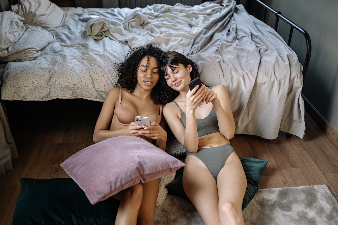 Two women in a cozy bedroom setting, enjoying time together and using their smartphones, embodying love and connection.